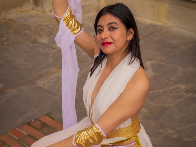 Samantha Smith, con cabello oscuro, lápiz labial rojo, vestido blanco, brazaletes dorados, sosteniendo un paño blanco, sonriendo, sentada, mostrando sus pechos, linda, sexy, piel de tono medio, segura de sí misma, atractiva..