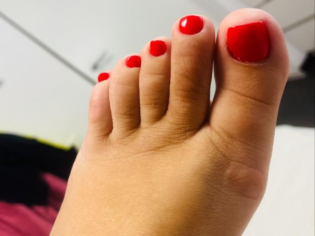 O pé de HotKrystall, com unhas dos pés vermelhas e brilhantes, dedos levemente curvados e pele bronzeada lisa. A foto se concentra em seu pé bem cuidado, mostrando suas unhas pintadas de vermelho em destaque..