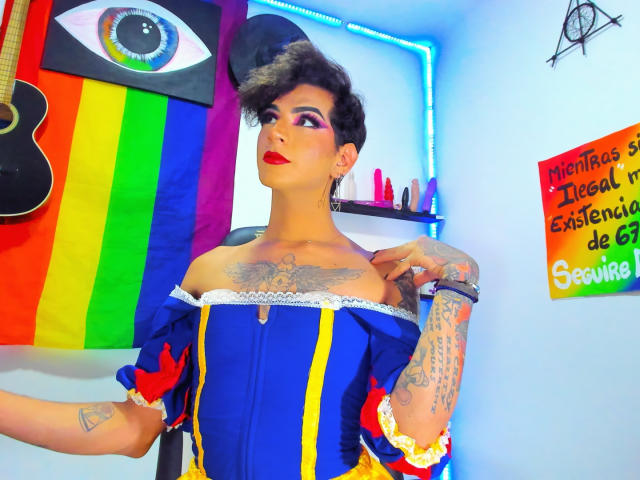 DesirTS com tatuagens, cabelo curto e escuro, batom vermelho, vestido azul e amarelo com um ombro de fora, levantando a mão, quarto colorido, bandeira arco-íris, luzes neon e brinquedos sexuais ao fundo..