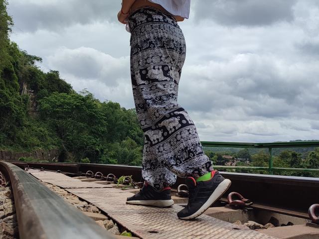 Les fesses de MelanyTessk, en pantalon noir et blanc de style bande dessinée, debout sur des rails de train rouillés, portant des baskets Adidas noires avec des chaussettes vert néon. Ciel nuageux, arbres verts en arrière-plan..