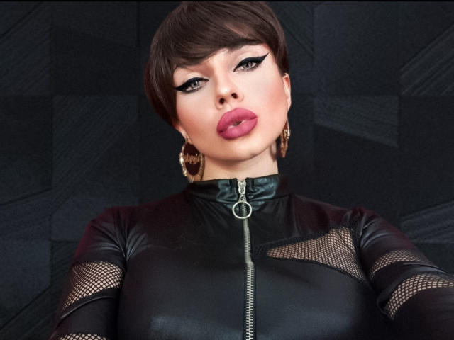 GoddessKaterina, com cabelo castanho curto, delineador preto, batom rosa e brincos de argola, vestindo uma blusa preta de couro com zíper e mangas de tela, mostrando seus seios..