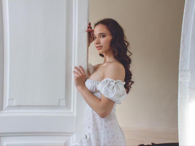 StacieLove mit langen, welligen, braunen Haaren, einem blumigen Kleid mit Schulterpartie, das eine weiße Tür hält, über die Schulter schaut, ein leichtes Lächeln, kleine Brüste, weiches Licht, elegant, steht im Türrahmen..