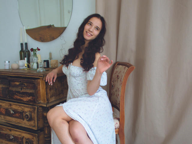 StacieLove, avec ses cheveux noirs bouclés, souriante, portant une robe blanche à une épaule, assise sur une chaise en bois, la main levée, avec sa poitrine visible, à côté d'une commode en bois avec des bouteilles et des bougies..
