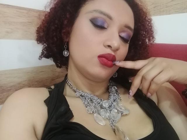 VioletaSwitch with curly red hair, purple eyeshadow, red lipstick, black top, silver necklace, and finger in mouth, showing off her tits and looking sexy.