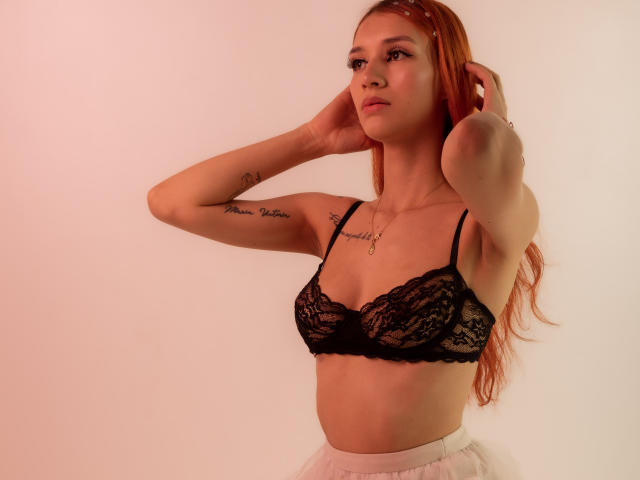 Greta Harris met rood haar, een zwarte kanten bh en een witte rok, handen in haar haar, tatoeages op haar arm, kijkt opzij, zachte roze verlichting..