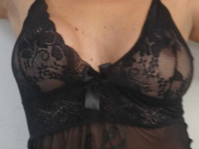 La lingerie in pizzo nero di NaomyHott69, trasparente e con motivi floreali, mostra i suoi seni attraverso il tessuto, con un piccolo fiocco nero al centro, braccia alzate, con la pelle visibile sopra..