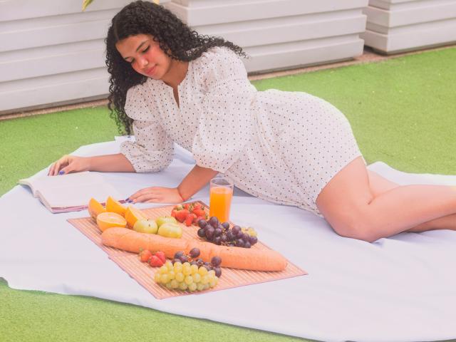 TerraFoxy, curly-haired, laying on white blanket, white polka-dot dress, eating, orange carrot, grapes, apple, orange, book, glass, green grass, relaxed, attractive, curvy, butt visible.