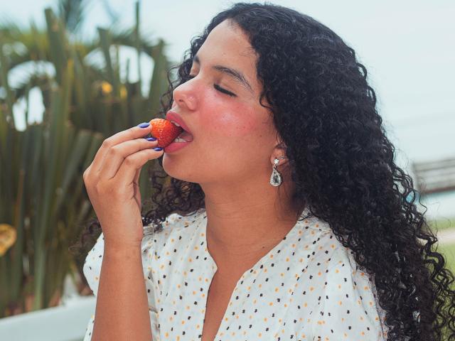 TerraFoxy with curly black hair, tan skin, and pink cheeks, eating a strawberry with her eyes closed. She's wearing a white polka dot shirt and dangling earrings.