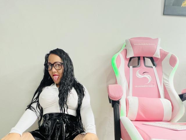 CarlaSexyFontaine, con cabello negro ondulado, gafas, lengua afuera, blusa blanca, falda de látex negra, sentada al lado de una silla de juego rosa y blanca. Los senos son de tamaño medio. La vulva no es visible..