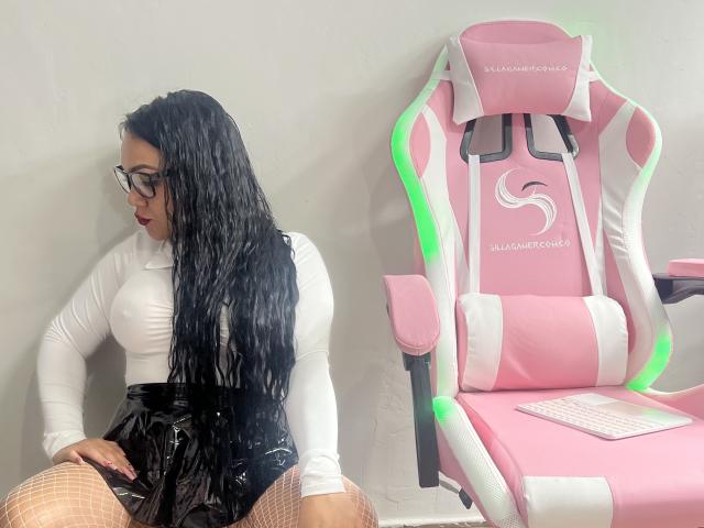 CarlaSexyFontaine met lang, nat zwart haar, een bril, een wit shirt met lange mouwen, een zwarte glanzende rok, netkousen, zittend naast een roze en witte gamingstoel met groene LED-verlichting..