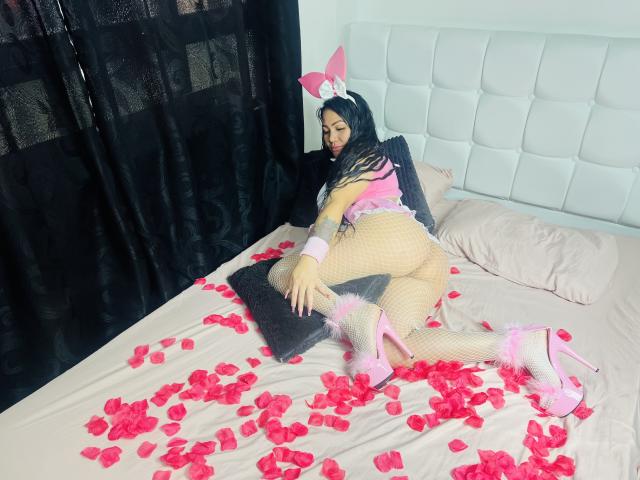 CarlaSexyFontaine with pink bunny ears, black lace top, fishnet stockings, pink heels, and pink fur trim, on a bed with rose petals, touching a black pillow.