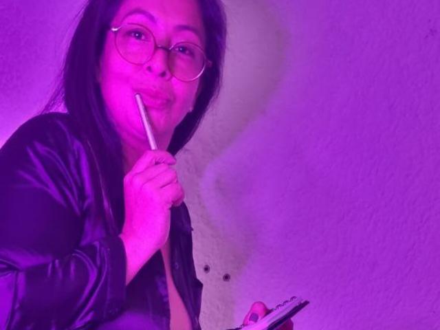 CatalinaJH, avec des lunettes roses, des cheveux noirs, tenant un crayon contre ses lèvres, portant une robe noire brillante, éclairée en violet, tenant un livret rouge et blanc dans sa main droite..
