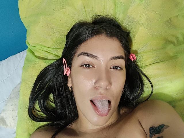 SharonKlart with black hair in pigtails, pink hair clips, nose ring, tongue out, glossy skin, small tattoo on right shoulder, lying on green pillow, blue wall in background.