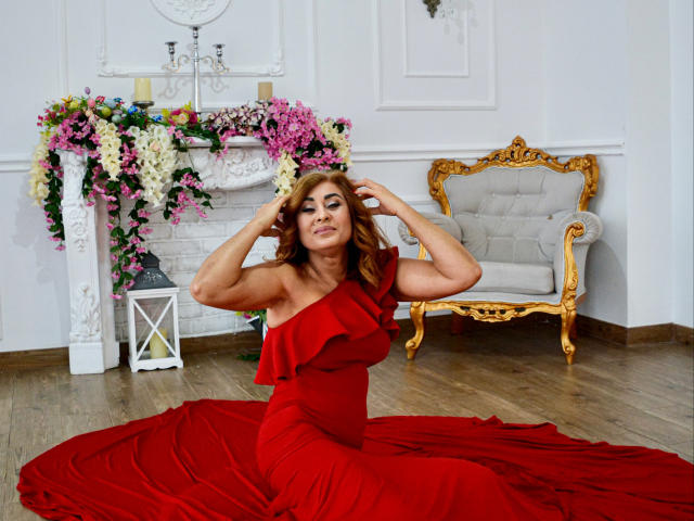 KarlaHartley in a red dress with ruffles, sitting on the floor, hands in her wavy brown hair, red dress pool, white and gold ornate chair in background.