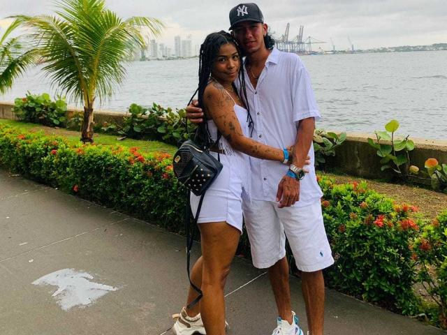MollyXDamiam with long black braids and tan skin, wearing white shorts and a black purse, stands with a tall guy in a NY cap and white clothes, by the water.