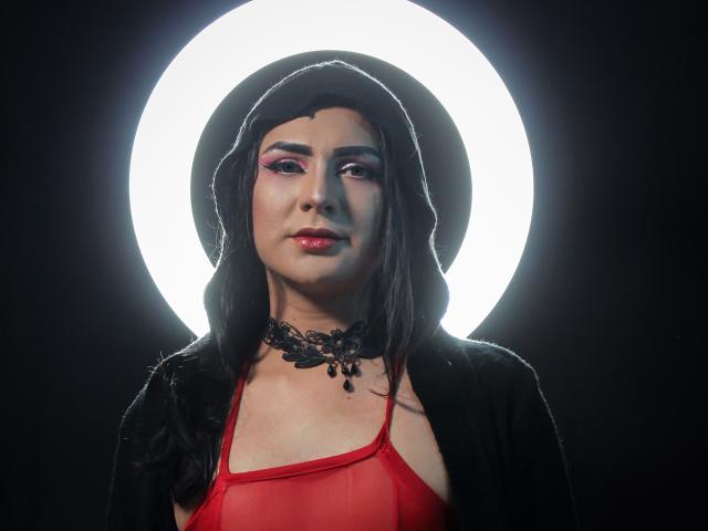 Iris Durand, met een rode doorschijnende top, een zwarte choker en een zwarte hoed, staat voor een heldere, ronde lamp. Ze heeft roze oogschaduw, rode lippenstift en een serieuze uitdrukking..