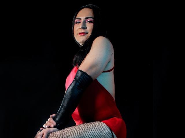 Iris Durand, con cabello negro largo, maquillaje rojo, vestido rojo, mangas negras en los brazos y medias de rejilla, sentada en un taburete, sonriendo, mostrando su trasero, sobre un fondo oscuro..