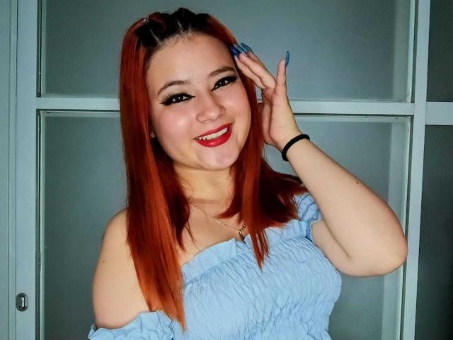 Venus: Capelli rossi, sorridente, top blu con spalline scoperte, mano tra i capelli, unghie blu, rossetto rosso, pelle chiara, fascia per capelli nera, vista frontale, seno di media grandezza, carina..