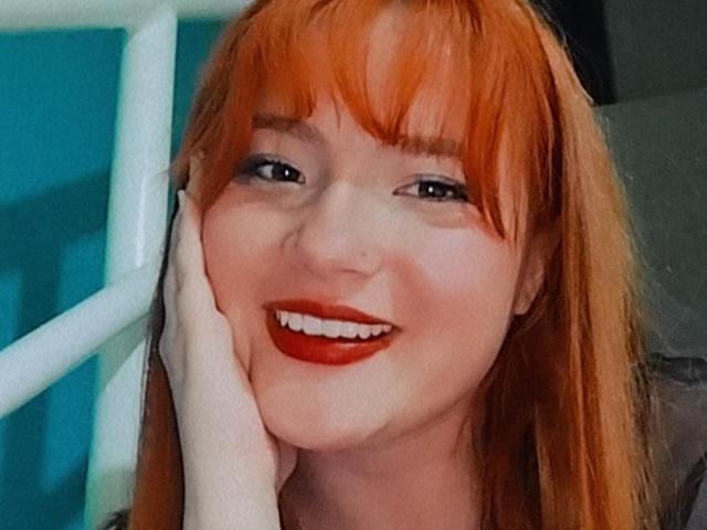 VenusCrazzy heeft fel oranje haar, een pony en ze glimlacht met rode lippenstift. Haar hand rust op haar wang. Ze draagt donkere oogmake-up. Haar tanden zijn zichtbaar..