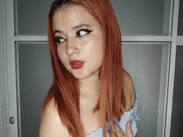 Venus: Impazzita, con i capelli rossi, un eyeliner nero spesso, rossetto rosso, che guarda di lato, indossa una maglietta azzurra a spalle scoperte, tenendola con la mano..