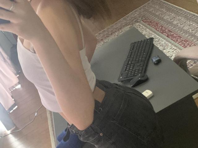 Tserera, con pechos pequeños, vestida con una camiseta blanca y pantalones vaqueros negros, de pie de lado a un escritorio con un teclado y un ratón, sosteniendo un teléfono en un selfie frente al espejo..