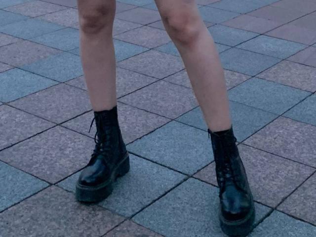 Tserera's legs in black, shiny, platform, lace-up boots, standing on a tiled, checker-patterned floor, showing pale skin and slightly bent knees.