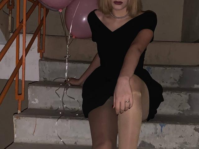 Tserera è seduta su delle scale sporche, indossa un vestito nero con una spalla scoperta, calze beige e rossetto scuro. A sinistra, un palloncino rosa con un filo d'argento, una collana choker e un anello alla mano destra..