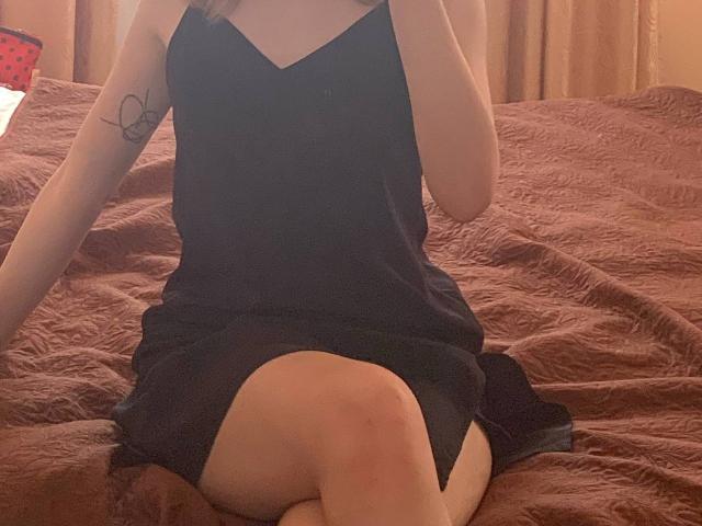 Tserera trägt ein schwarzes Kleid und sitzt auf einem braunen Bett, die Beine gekreuzt, wobei ihr Hintern zu sehen ist. Sie hat ein Schmetterlings-Tattoo auf ihrem rechten Arm und hält ihr Handy..