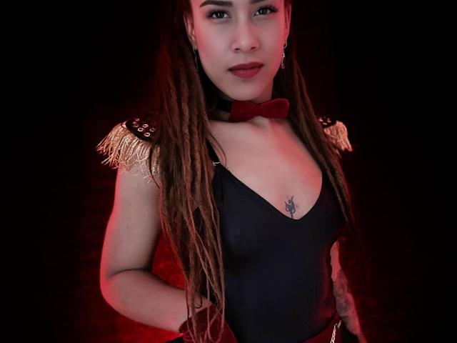 Kendall Quinn, com longos dreadlocks castanhos, vestindo um collant preto, um laço vermelho e ombreiras douradas, tem uma pequena tatuagem abaixo dos seios e um sorriso adorável..