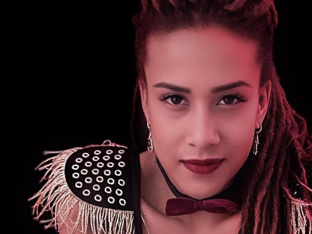 KendallQuinn has dark red dreadlocks, wearing a spiked shoulder epaulet, black choker with red bow, and dark lipstick, with a red light glow on her face.