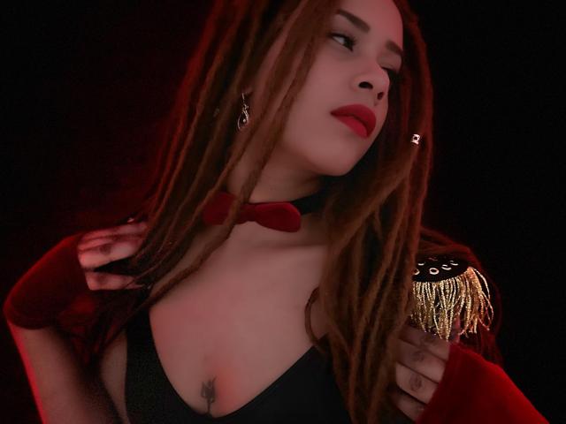 KendallQuinn with long brown dreads, red lips, black choker, red bowtie, and red jacket, showing off gold epaulets and black cleavage, holding jacket, red light.
