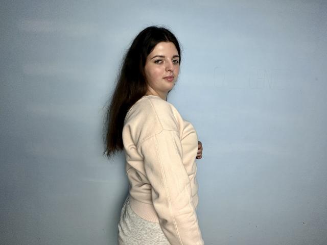 PamDares, con cabello castaño largo, piel pálida y senos pequeños, vestida con una sudadera beige y pantalones blancos, está de pie de lado contra una pared azul claro, mirando a la cámara..