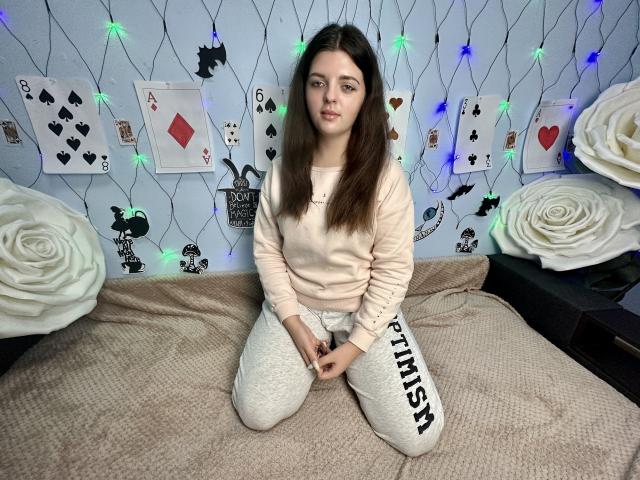PamDares, met lang bruin haar, zit op een bed in een roze trui en witte broek met de tekst "PIMPS", met haar handen in elkaar gevouwen, en kijkt naar de camera. Er hangen blauwe en groene lichtslingers..