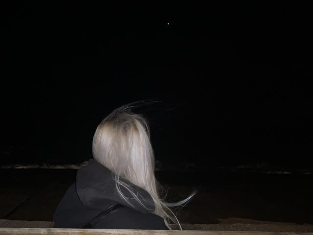 Tserera, con el pelo largo y blanco, vestida con una sudadera negra, mirando hacia un pequeño punto brillante en el oscuro cielo nocturno..