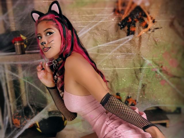 Aby Coleman, avec des cheveux rose et noir, des oreilles de chat, des bras en résille noire, une robe rose, un collier et du maquillage imitant des moustaches, tenant des lunettes, regardant par-dessus son épaule, dans une pièce effrayante recouverte de toiles d'araignées..