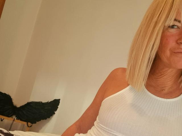 Une femme blonde, BlackAngeel, avec une coupe au carré, portant un débardeur blanc côtelé, qui révèle ses petites seins et une légère démarcation de bronzage, dans une pièce légèrement éclairée..