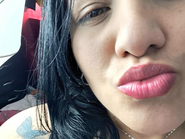 Het gezicht van Viviana Thompson, met zwart haar, glanzende roze lippen en een tatoeage van een blauwe vlinder op haar schouder. Haar neus is iets omhoog gekeerd, en ze draagt een dunne ketting..