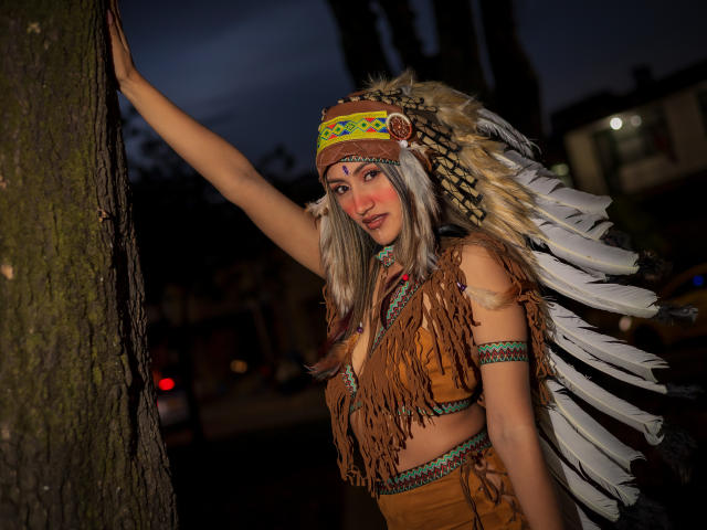 Helen Madisson, avec ses longs cheveux gris, portant un haut marron à franges, un bandeau coloré orné de perles et une coiffe blanche ornée de plumes, appuyée contre un arbre la nuit, son bras levé..