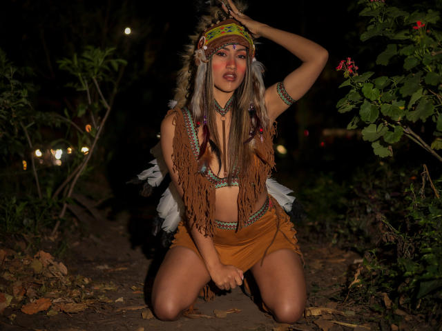 HelenMadisson kneels on dirt, wearing a fringed brown top and shorts, with a colorful headband, arm bangle, and feathered back. Her tits are visible, dark night background.