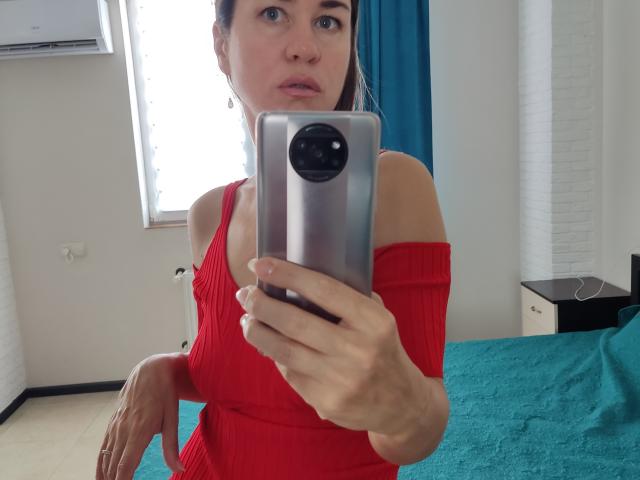 Karamel69, con gli occhi azzurri, indossa una maglia a coste rossa con una spalla scoperta, sta scattando un selfie allo specchio con uno smartphone argentato, mostrando il seno e appare sorpresa..