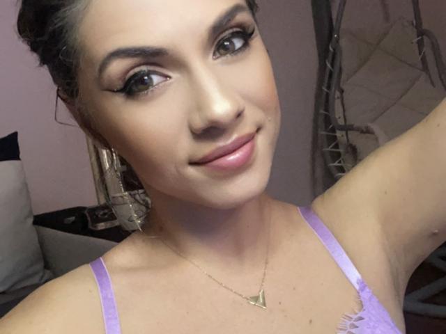 LucilleXSugar tiene la piel de color marrón claro, el cabello castaño oscuro recogido en un moño, y lleva un sujetador de encaje morado con aretes dorados y un collar triangular. Está sonriendo y lleva delineador de ojos..