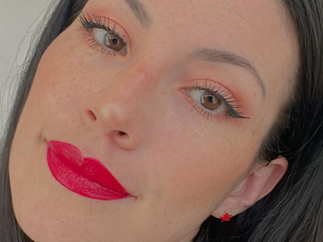 Liliysweet's face with red lipstick, brown eyes, black eyeliner, orange eyeshadow, black eyebrows, and red butterfly earrings, showing a slight smile. Her skin is fair with freckles.