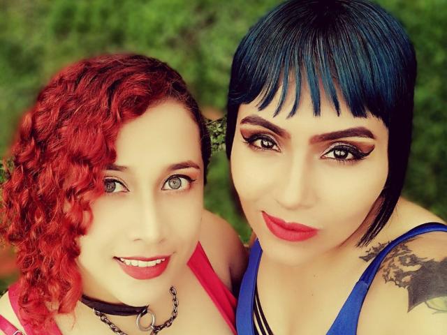 AbbyElecktra met krullend rood haar en groene ogen, gekleed in een roze top en een zwarte choker, naast AbbyElecktra met kort blauw haar, rode lippenstift en een blauwe top, die een tatoeage laat zien..