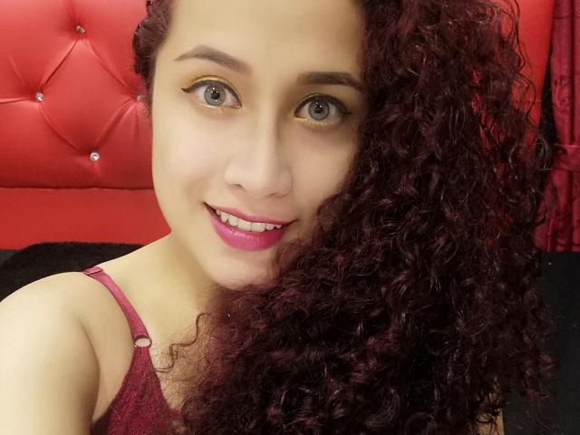 AbbyElecktra, con i capelli rossi ricci, la pelle chiara, il rossetto rosa e l'eyeliner giallo, sorride alla fotocamera, indossando una camicetta rossa in pizzo che mostra leggermente il seno..