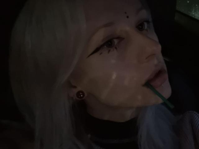 ExcitedBlizzard has pale skin, white hair, black eyeliner, small black moles on her face, and a green straw in her mouth, with dark shadows and dim lighting.