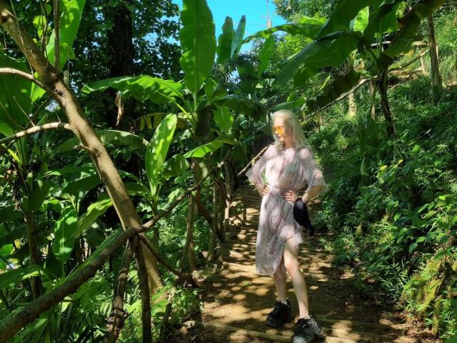 ExcitedBlizzard stands in a lush jungle, wearing a sheer, white, patterned dress with a thigh slit, exposing her bare, white, hairy-ass. She's holding a black bag.