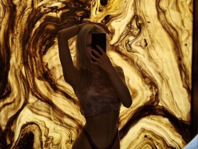 ExcitedBlizzard with blonde hair, arms up, wearing a tie-dye crop top and black panties, holding a phone, covering her face, against a marbled brown and gold background.
