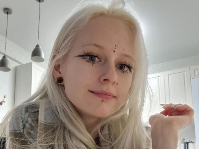 ExcitedBlizzard has pale skin, blonde hair, black eye makeup, and black ear gauges, with a small nose piercing and lip piercing, smiling at the camera.