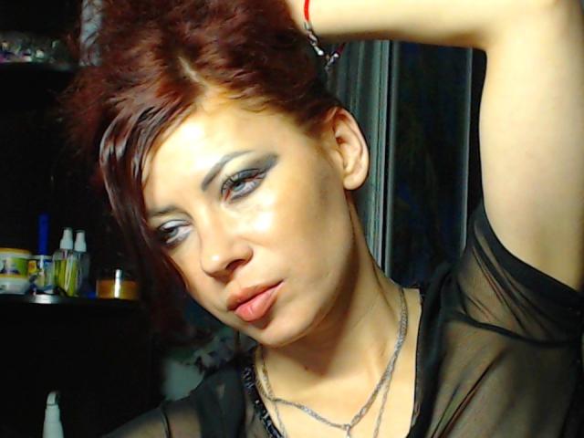 JuliaIce tem cabelo ruivo, delineador preto e uma expressão séria, vestindo uma blusa preta transparente, com um braço levantado e um colar, olhando para baixo com um leve semblante de descontentamento..