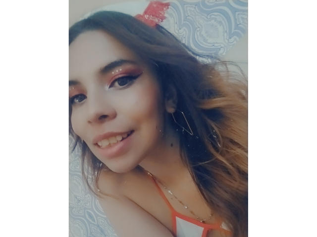 VioletaStone has wavy brown hair, orange eyeshadow, and a cute smile with a slightly open mouth, showing her small, shiny teeth. She's wearing a red top.